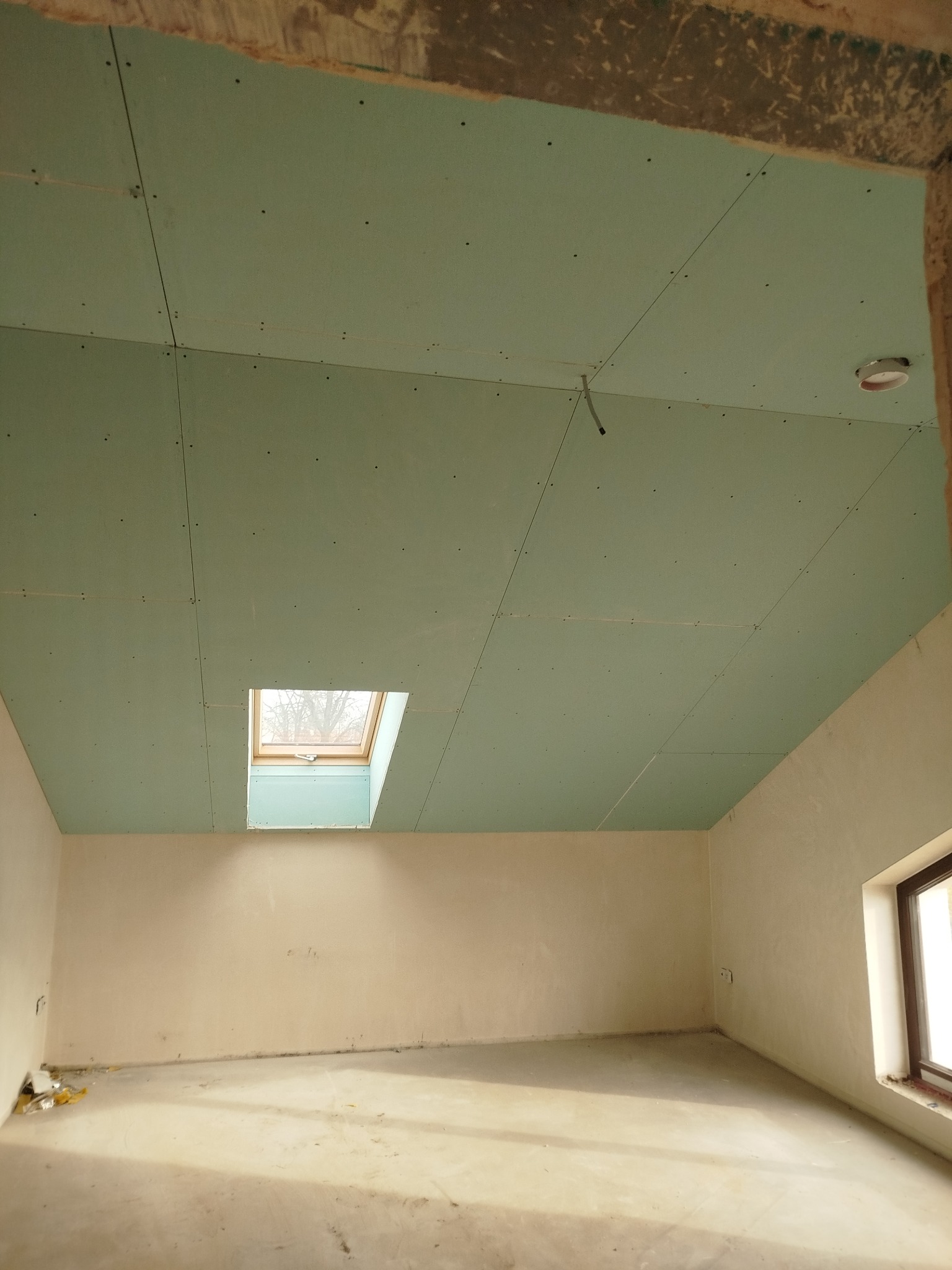 Pokój w trakcie remontu: zielone płyty karton-gips na suficie z oknem dachowym, surowe ściany i podłoga. Widoczny fragment okna w ścianie bocznej.