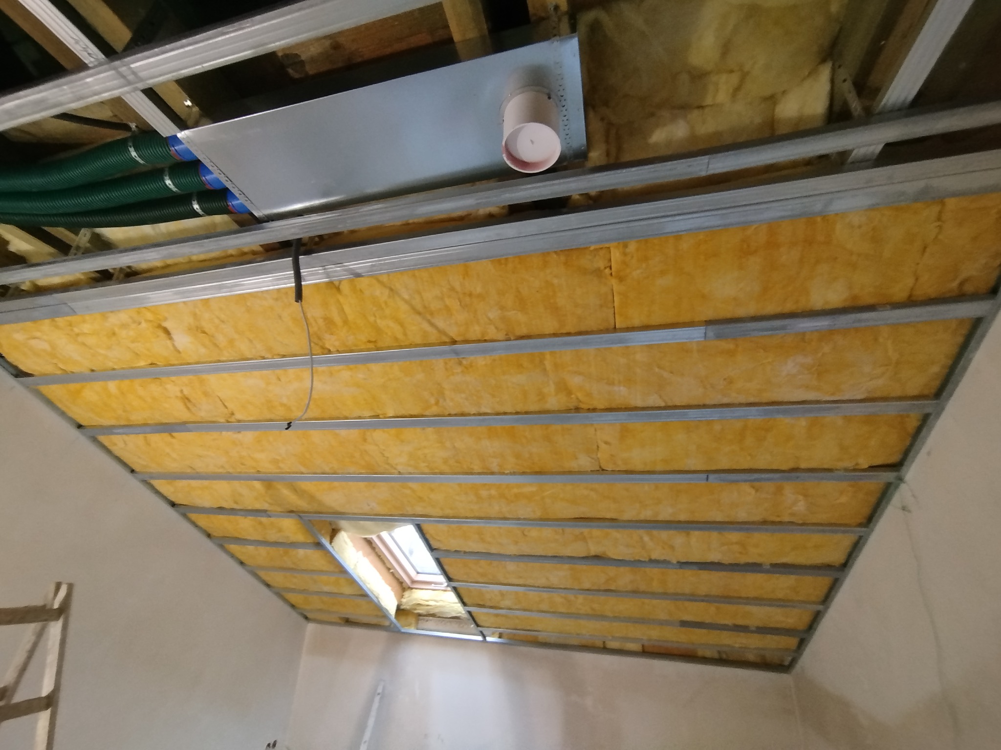 Montaż izolacji z wełny mineralnej w metalowej konstrukcji sufitu podwieszanego z widocznym oknem dachowym i elementami wentylacji.