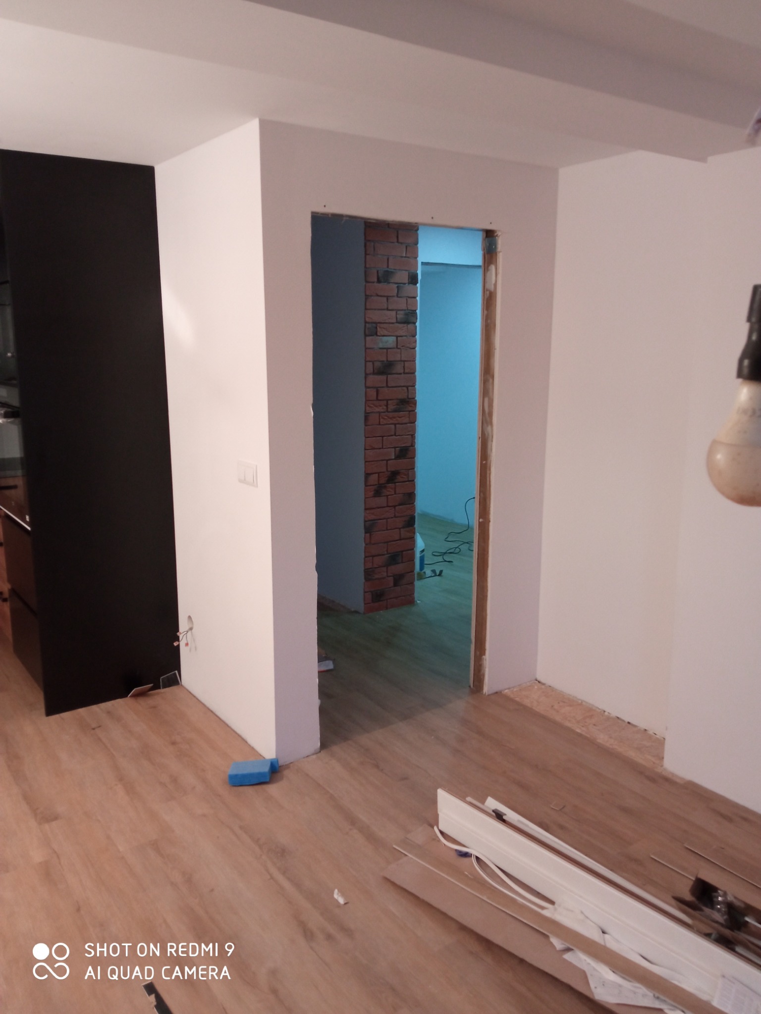 Wnętrze w trakcie remontu: położone panele podłogowe, widoczne listwy przypodłogowe, ściany pomalowane na biało, fragment ściany z cegły w przejściu.