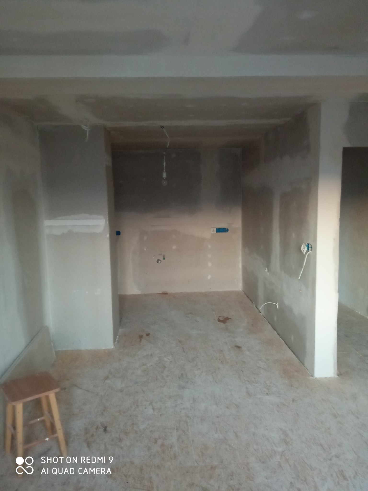 Wnętrze w trakcie szpachlowania: ściany pokryte gładzią, widoczne przewody elektryczne, drewniany stołek w rogu. Etap prac wykończeniowych przed malowaniem.