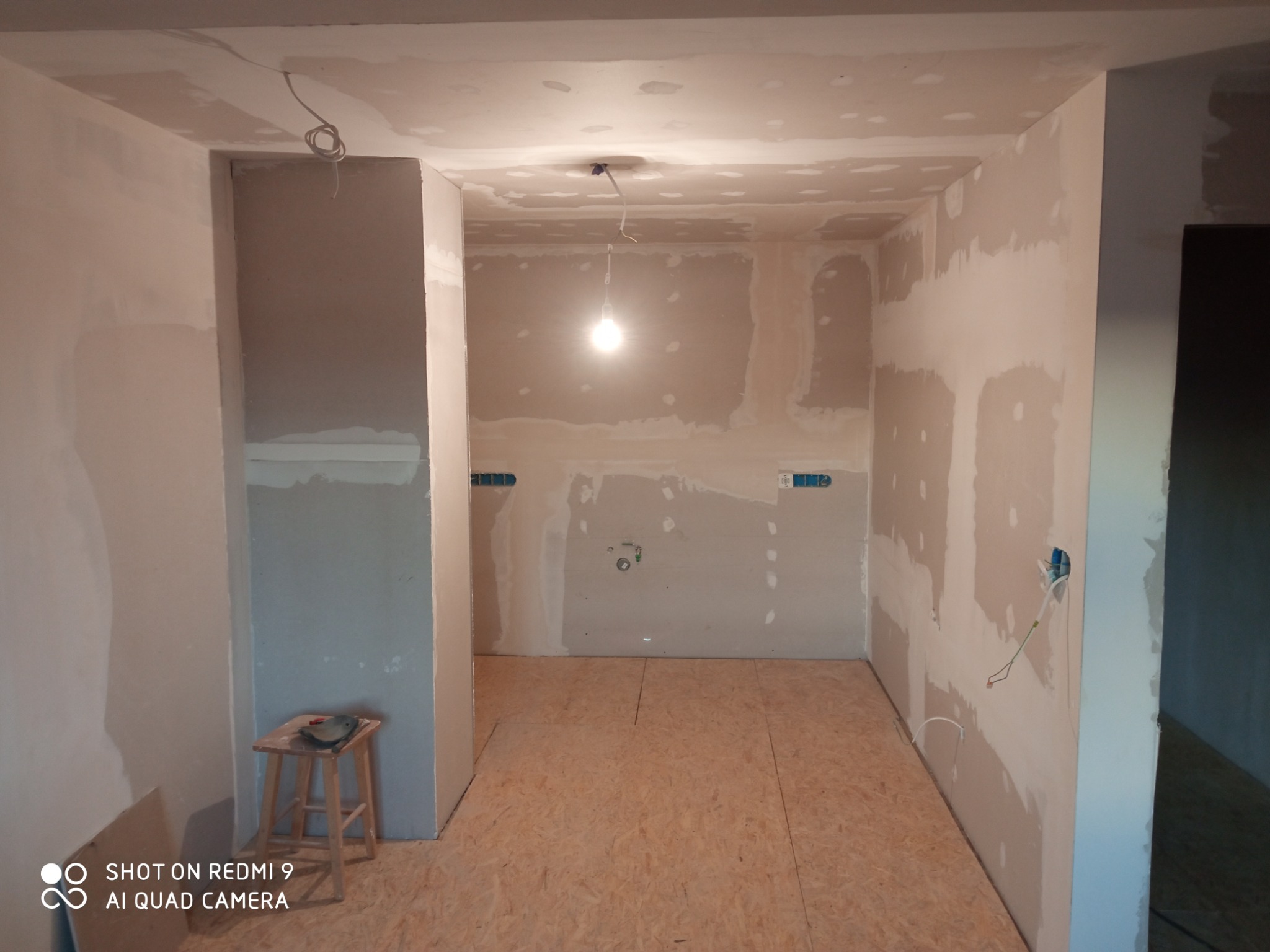 Wnętrze w trakcie prac: ściany z płyt gipsowo-kartonowych, częściowo wyszpachlowane, podłoga z płyt OSB, widoczne instalacje elektryczne i lampa wisząca.