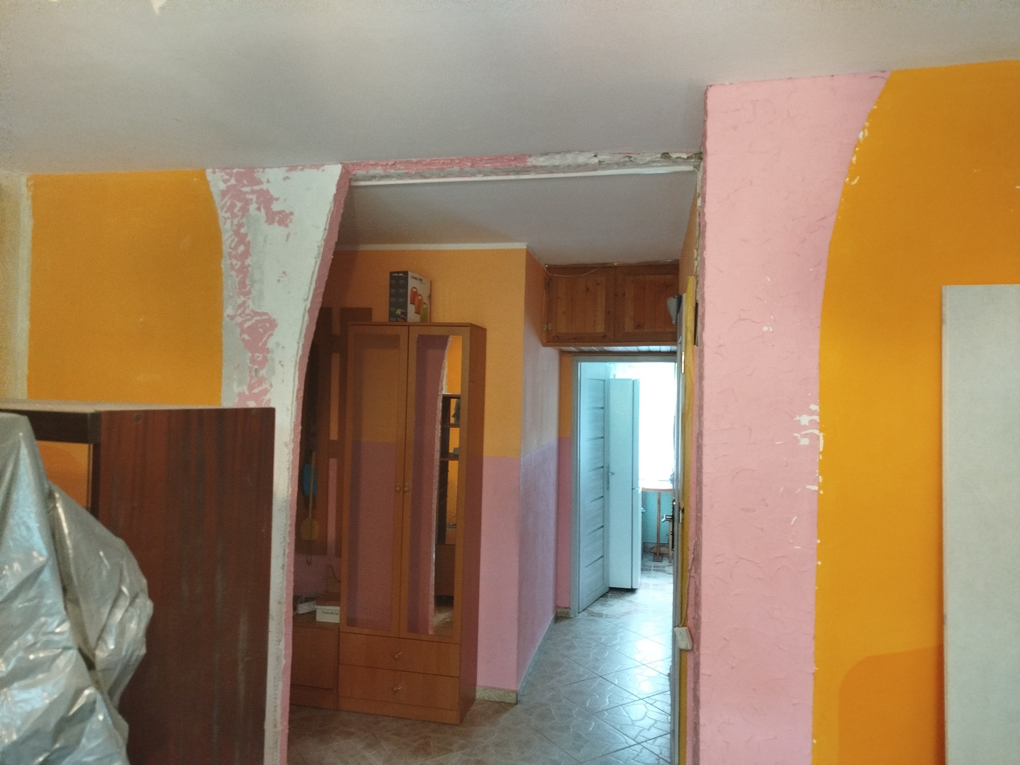 Pokój w trakcie remontu: fragment pomarańczowej ściany z odchodzącą farbą, różowy filar i widok na dalsze pomieszczenia. Widoczny mebel częściowo zakryty folią.
