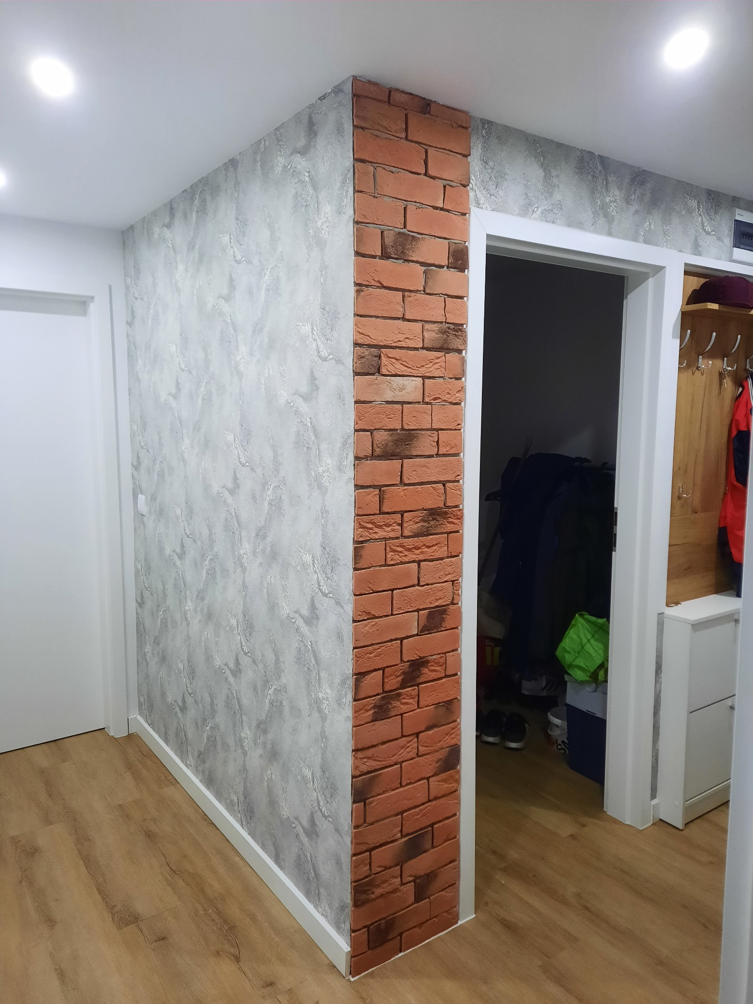 Korytarz po remoncie: ściana z efektem betonu, narożnik wykończony cegłą dekoracyjną. Widoczne białe drzwi i fragment podłogi z paneli. Oświetlenie sufitowe.