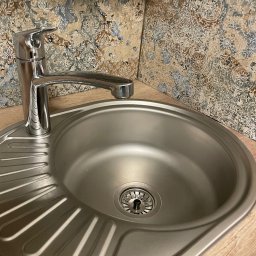 Amadin Cleaning Solution Oleksii Zazymko - Błyszczący, stalowy zlew kuchenny z nowoczesną baterią na tle dekoracyjnych płytek ściennych w odcieniach beżu i błękitu.