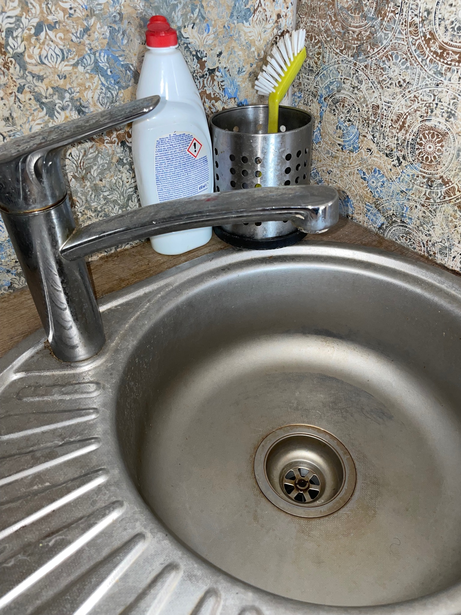 Metalowy zlew kuchenny z kranem, płynem do naczyń i szczotką na tle wzorzystych płytek.