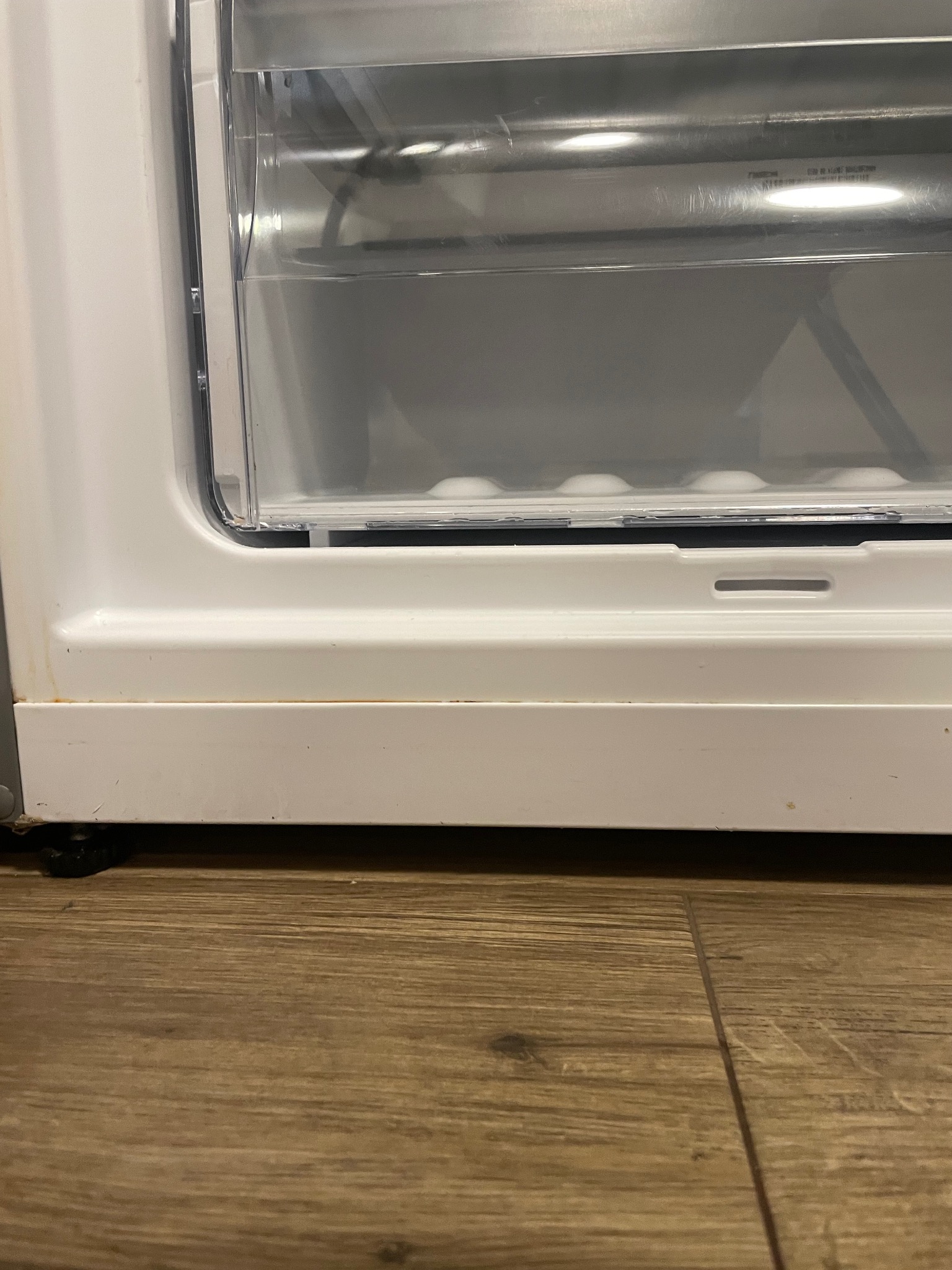 Dolna część białej lodówki z widocznymi zabrudzeniami i plamami na plastikowej obudowie, stojąca na drewnianej podłodze.