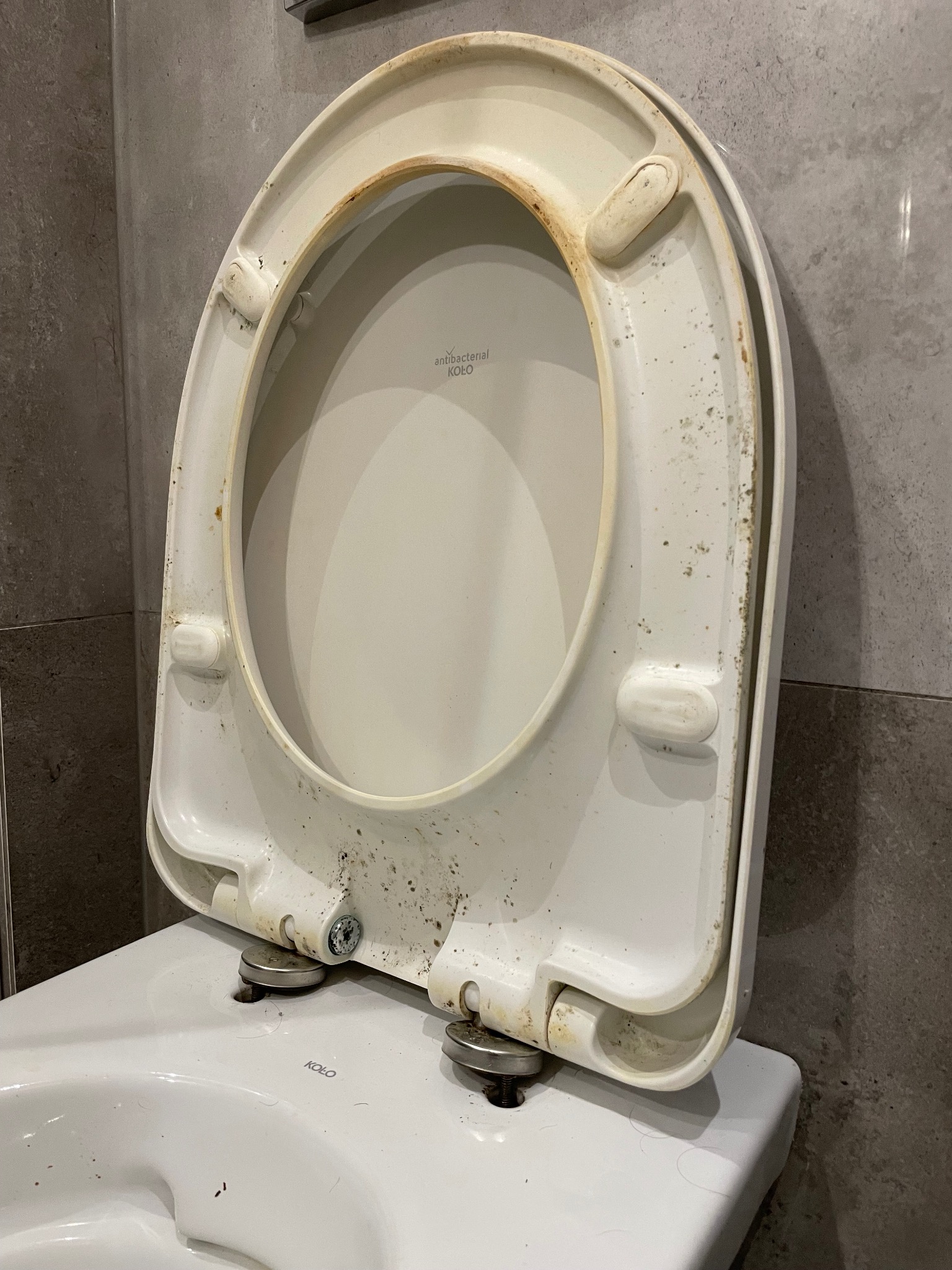 Brudna, biała deska sedesowa marki Koło z widocznymi śladami rdzy i zabrudzeniami, otwarta i oparta o ścianę z szarych płytek.