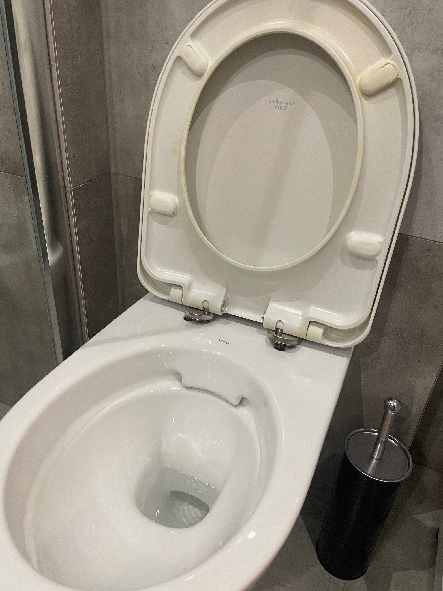 Ceramiczna toaleta marki Koło z otwartą klapą, widoczne ślady użytkowania i szczotka do WC w czarnym pojemniku na tle szarych płytek.
