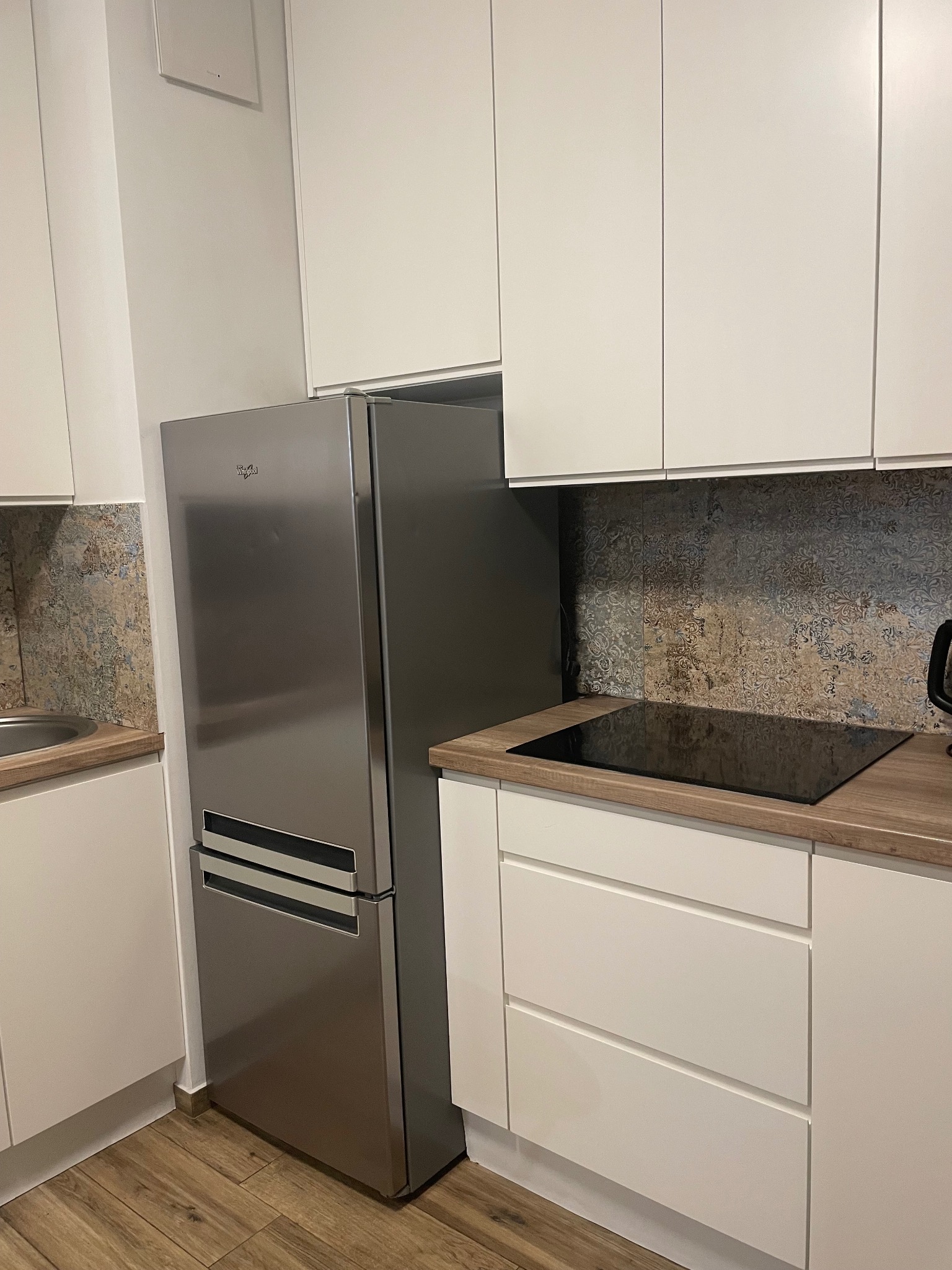 Nowoczesna kuchnia z białymi szafkami, drewnianym blatem i metalową lodówką Whirlpool, na podłodze brązowe panele.