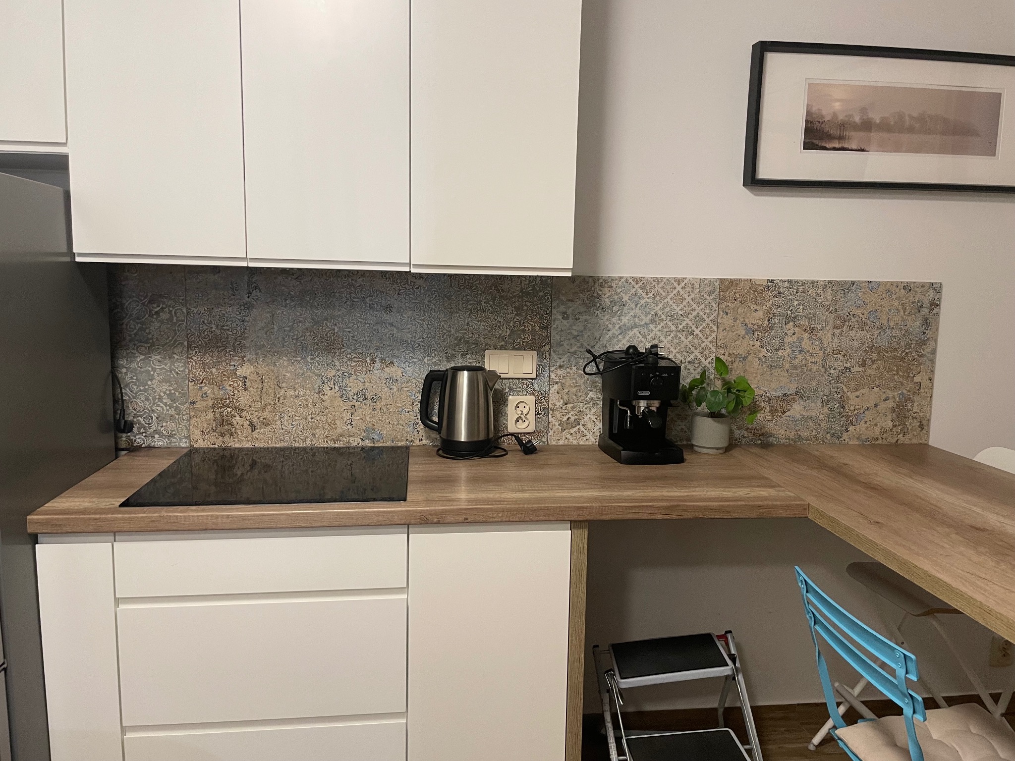 Nowoczesna kuchnia z białymi szafkami, drewnianym blatem, wzorzystą ścianą nad blatem, ekspresem do kawy, czajnikiem elektrycznym i obrazem na ścianie.