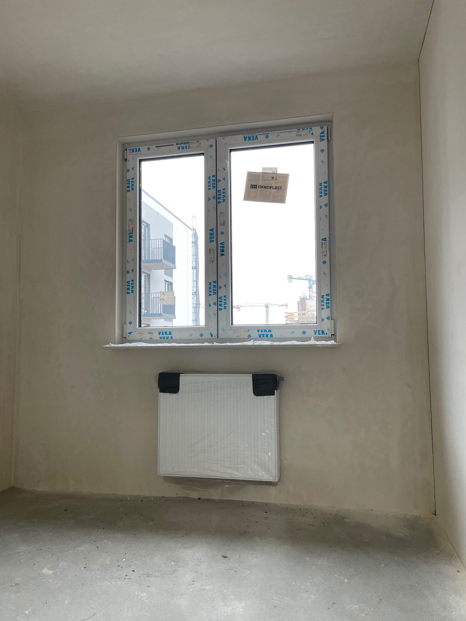 Surowe wnętrze w stanie deweloperskim z nowym oknem PCV z naklejkami producenta i grzejnikiem pod oknem, widok na budowę za oknem.