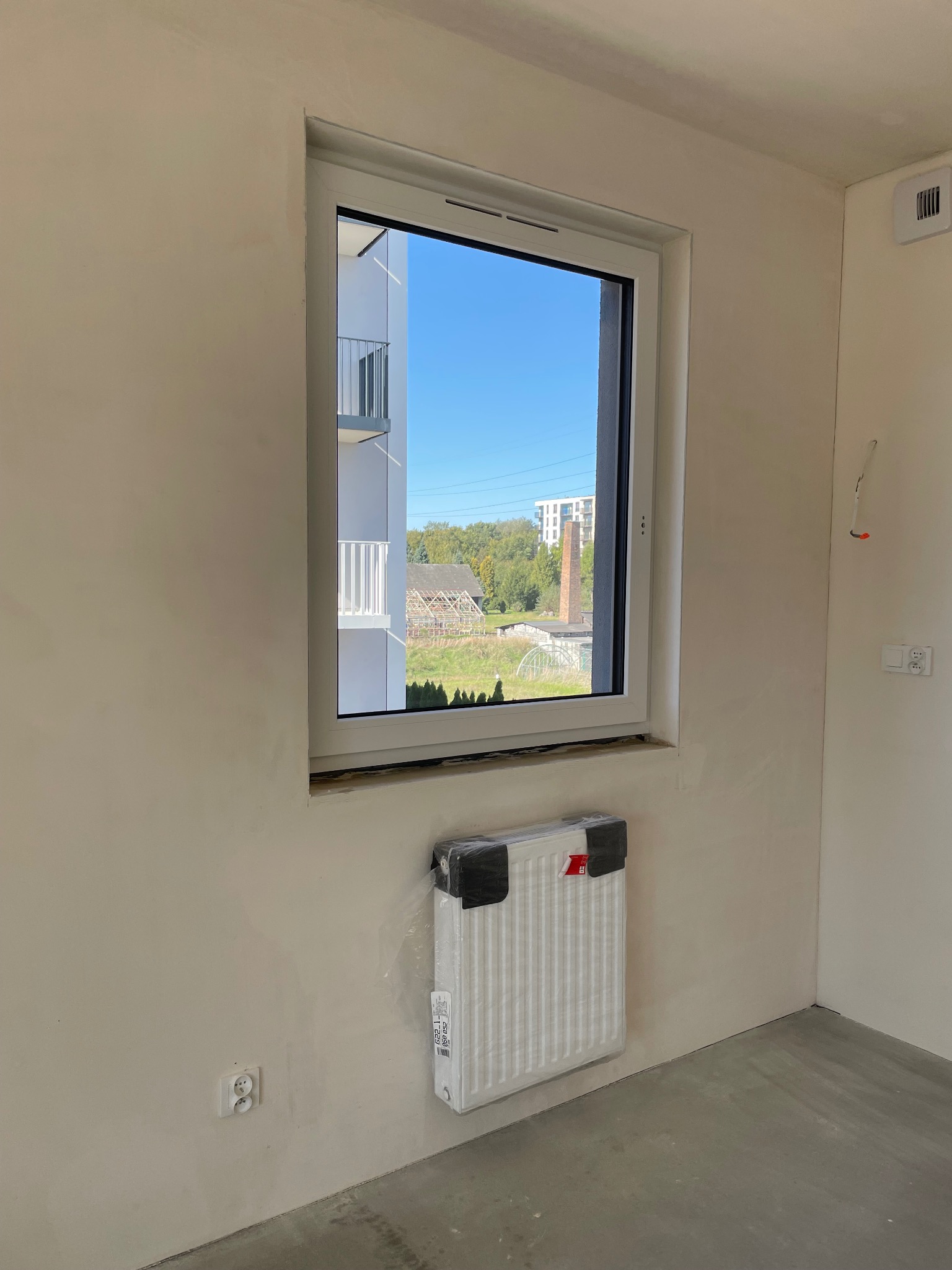 Nowe okno PCV z widokiem na budynek mieszkalny i komin fabryczny w tle, grzejnik w folii ochronnej pod oknem, ściany po gładzi, surowa podłoga.