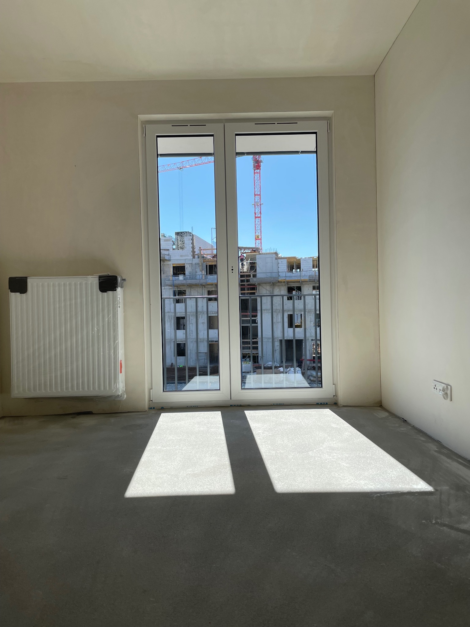 Puste, jasne pomieszczenie z surowymi ścianami i podłogą, okno balkonowe z widokiem na budowę z żurawiem, grzejnik w folii ochronnej po lewej.