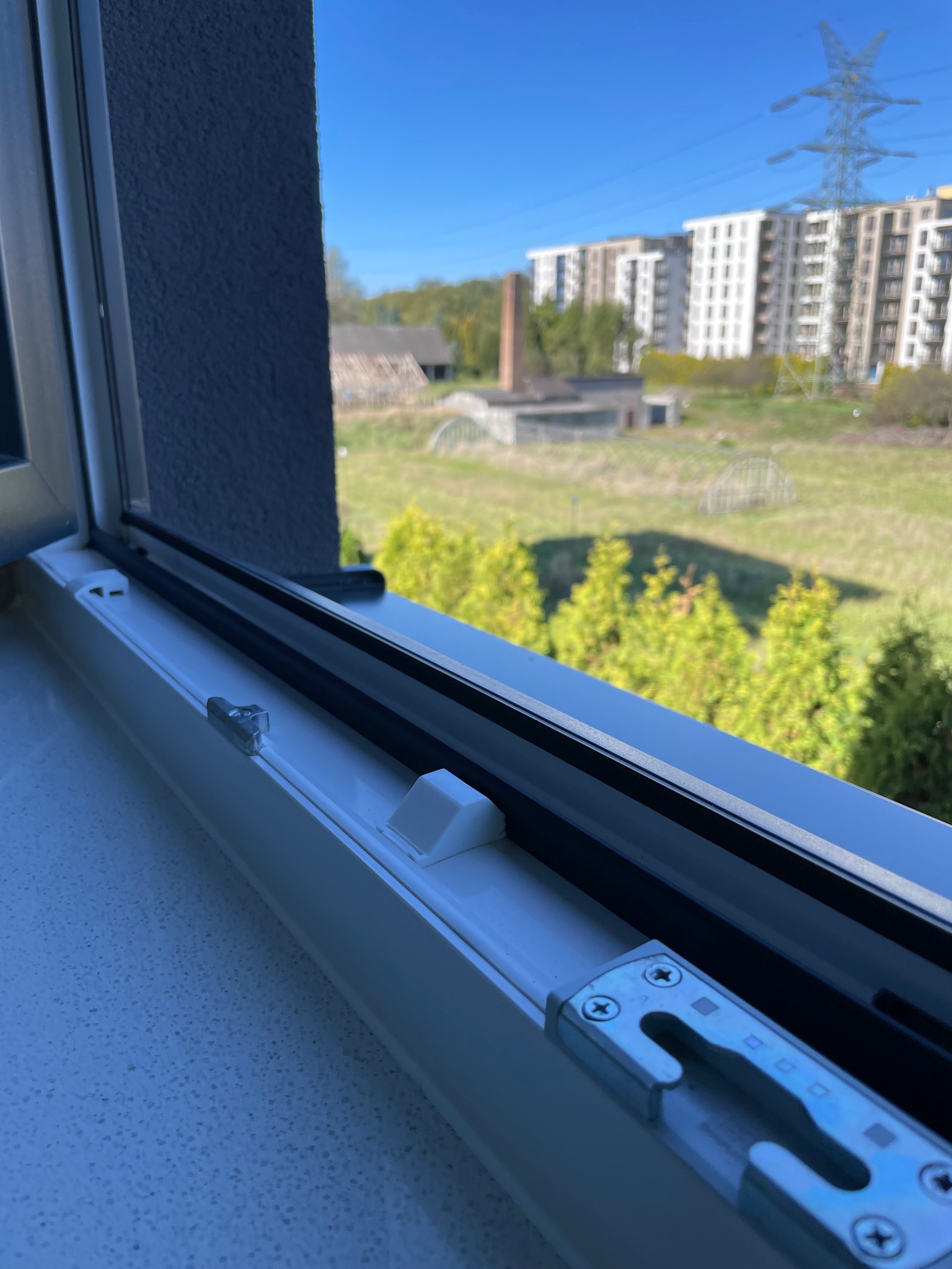 Detal widoku z okna PCV na zewnątrz: zabudowania gospodarcze, drzewa iglaste i blok mieszkalny z wieżą wysokiego napięcia w tle.