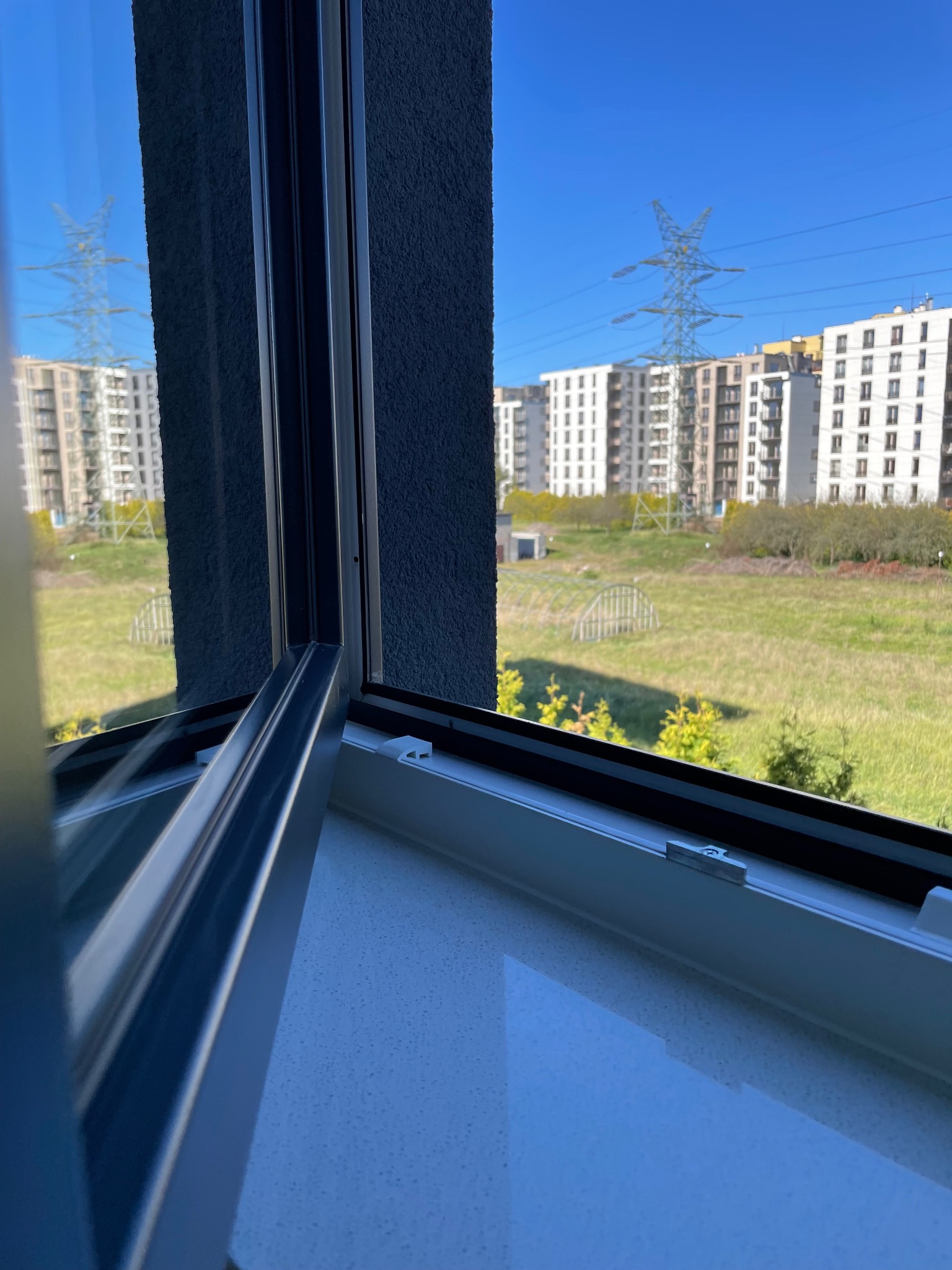 Otwarty fragment okna z widokiem na osiedle mieszkaniowe, trawnik i słup wysokiego napięcia w słoneczny dzień.