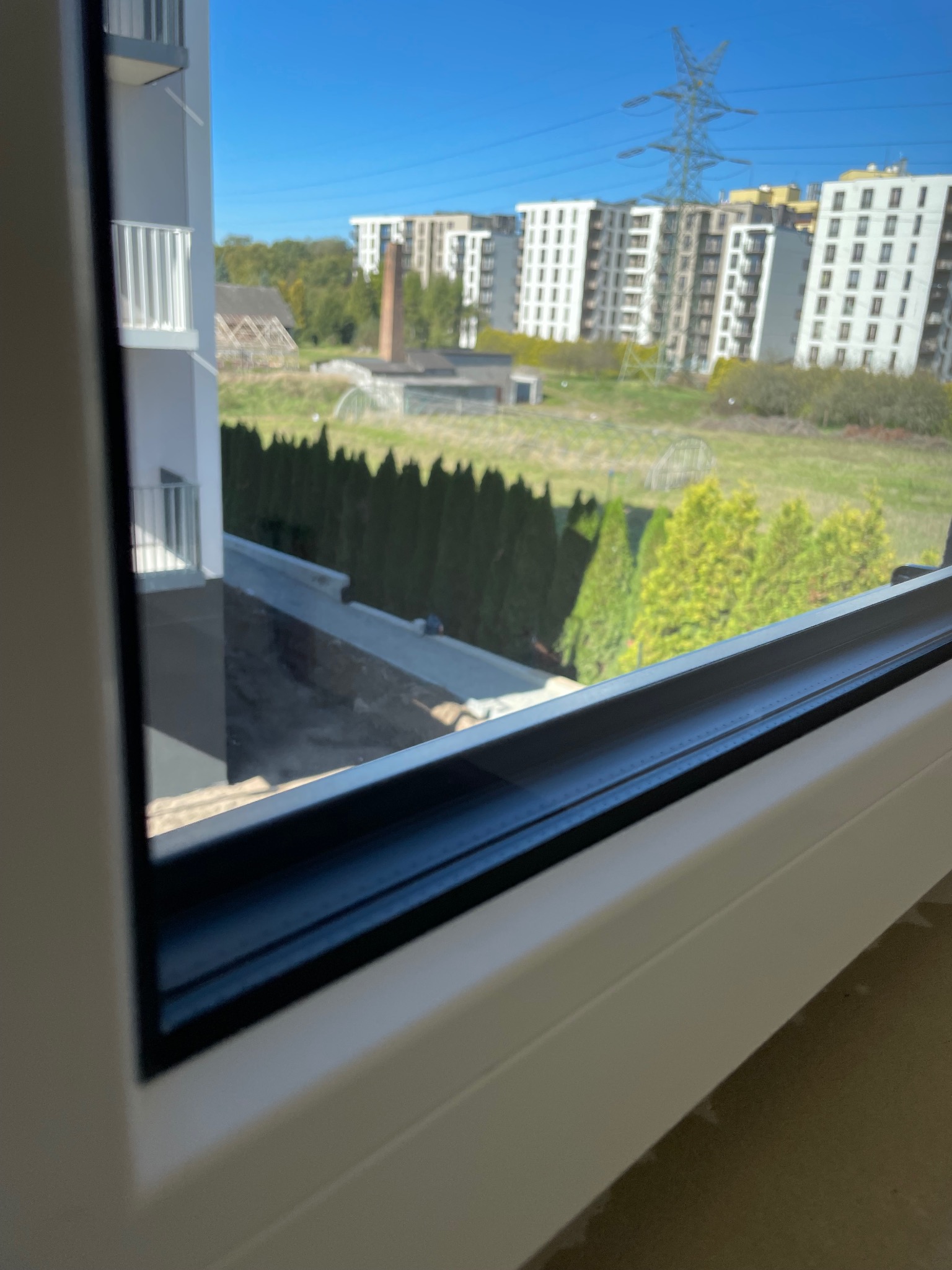 Widok z okna na osiedle z zielenią, widoczne budynki mieszkalne, drzewa i linia wysokiego napięcia.