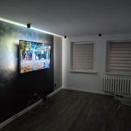 Balticconstruction - Nowoczesny salon z ciemną ścianą akcentującą, na której wisi telewizor, drewniana podłoga, białe ściany, grzejnik pod oknem i szklany stolik kawowy.