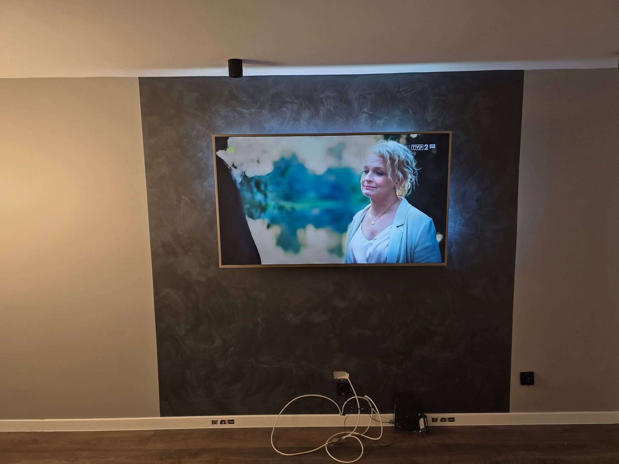 Telewizor zawieszony na ścianie z dekoracyjnym szpachlowaniem w odcieniach szarości, widoczne kable i gniazdka przy podłodze.