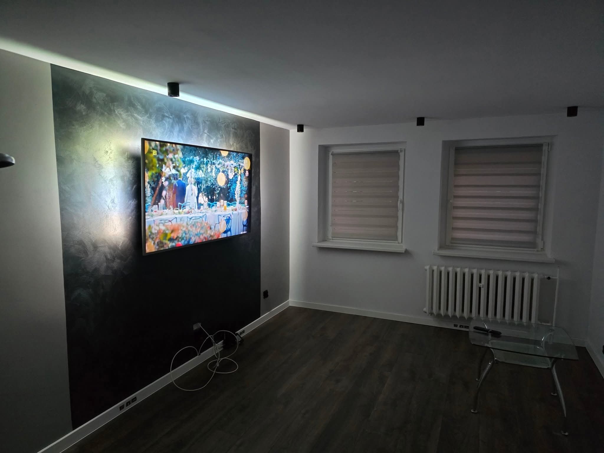 Nowoczesny salon z ciemną ścianą akcentującą, na której wisi telewizor, drewniana podłoga, białe ściany, grzejnik pod oknem i szklany stolik kawowy.