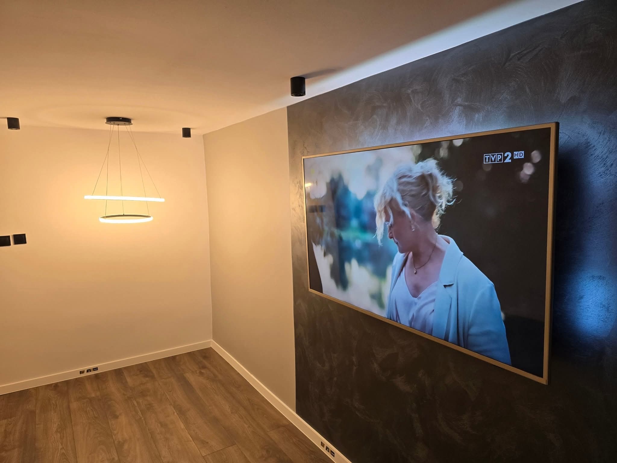 Nowoczesny salon z drewnianą podłogą, ścianą z efektem dekoracyjnym, podwieszanym oświetleniem LED i telewizorem z cienką ramką.