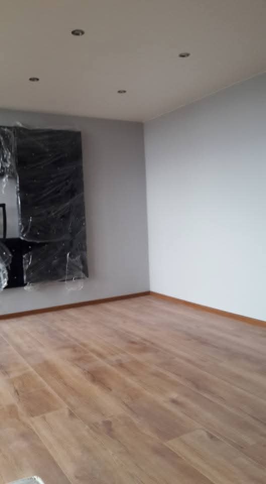 Pusty pokój z nową, laminowaną podłogą, świeżo pomalowanymi ścianami i sufitem z wpuszczonymi lampami, mebel zabezpieczony folią ochronną przy ścianie.