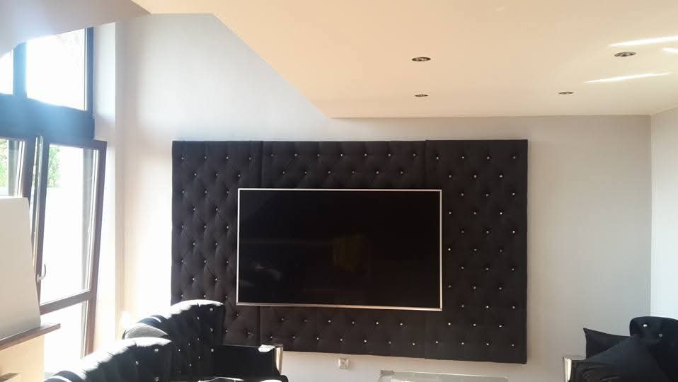 Telewizor zawieszony na ścianie z czarnym, pikowanym panelem tapicerowanym z ozdobnymi guzikami, w jasnym salonie z dużym oknem.