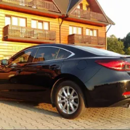 Czarny samochód Mazda 6 zaparkowany przed drewnianym budynkiem z balkonami, widok z tyłu i z boku, w tle drugi samochód.