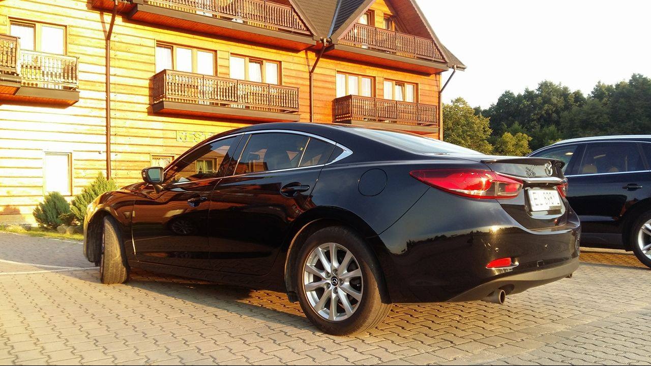 Czarny samochód Mazda 6 zaparkowany przed drewnianym budynkiem z balkonami, widok z tyłu i z boku, w tle drugi samochód.