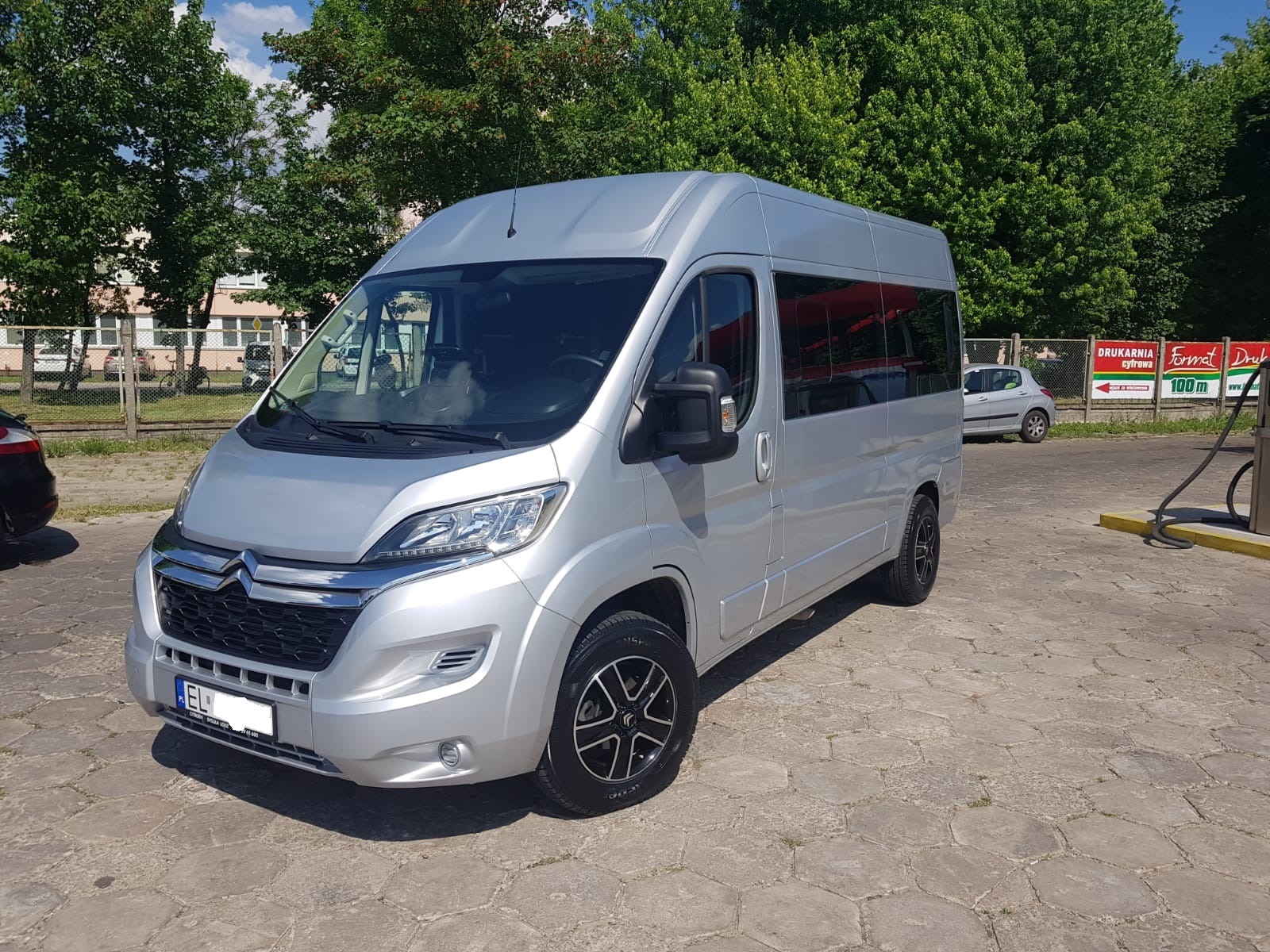 Srebrny bus Citroen Jumper z czarnymi felgami, zaparkowany na brukowanym placu w słoneczny dzień, widoczne reklamy drukarni w tle.