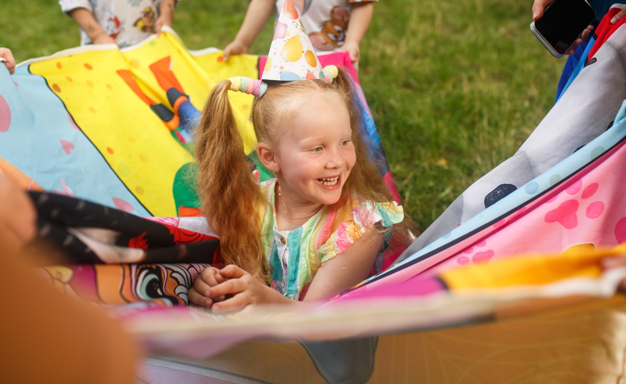 Uśmiechnięta dziewczynka w czapce urodzinowej, bawiąca się kolorową chustą animacyjną z innymi dziećmi na trawie podczas imprezy plenerowej.