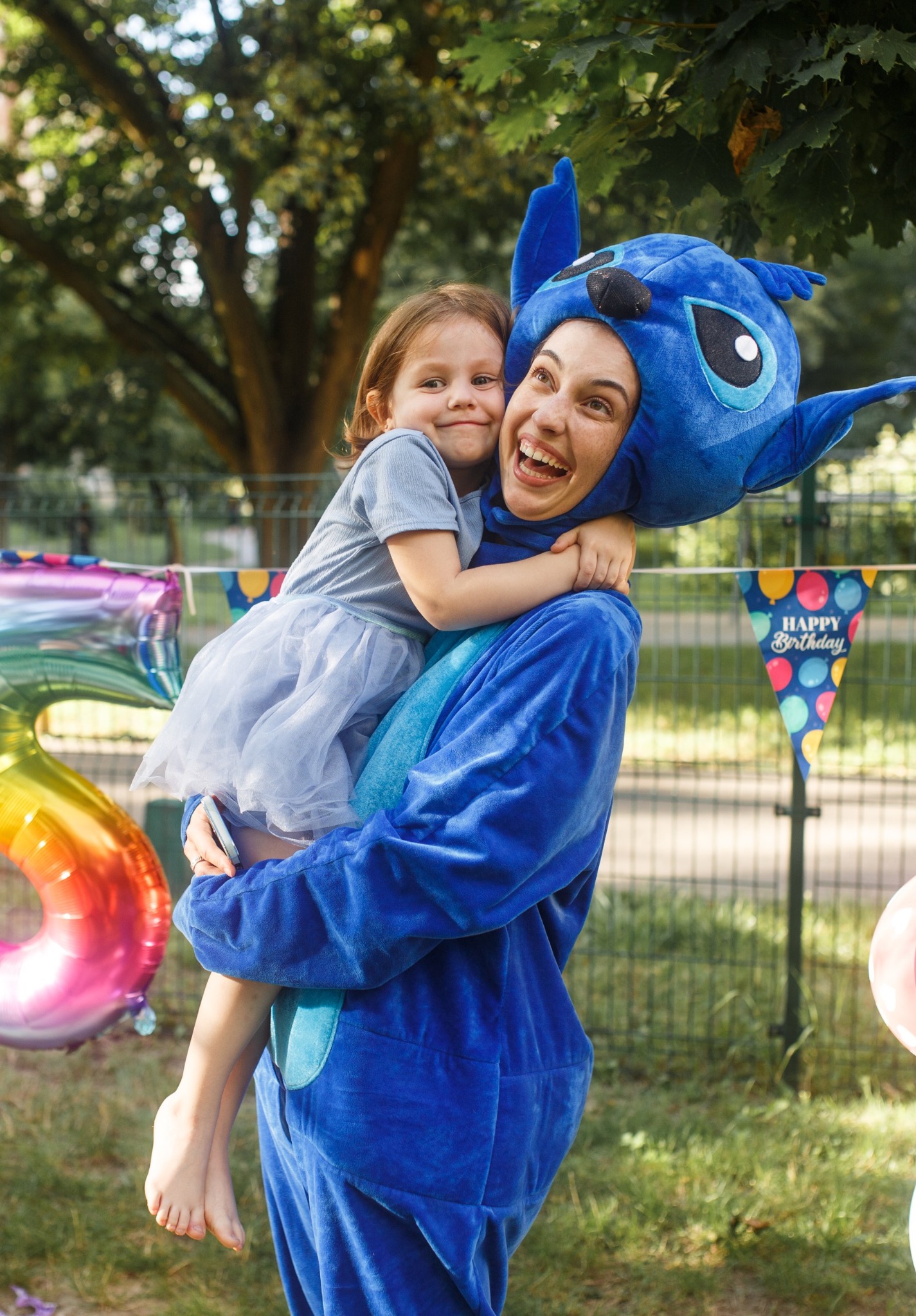 Animator w kostiumie Stitch trzyma uśmiechniętą dziewczynkę na tle dekoracji urodzinowych, w parku. Radosna atmosfera.