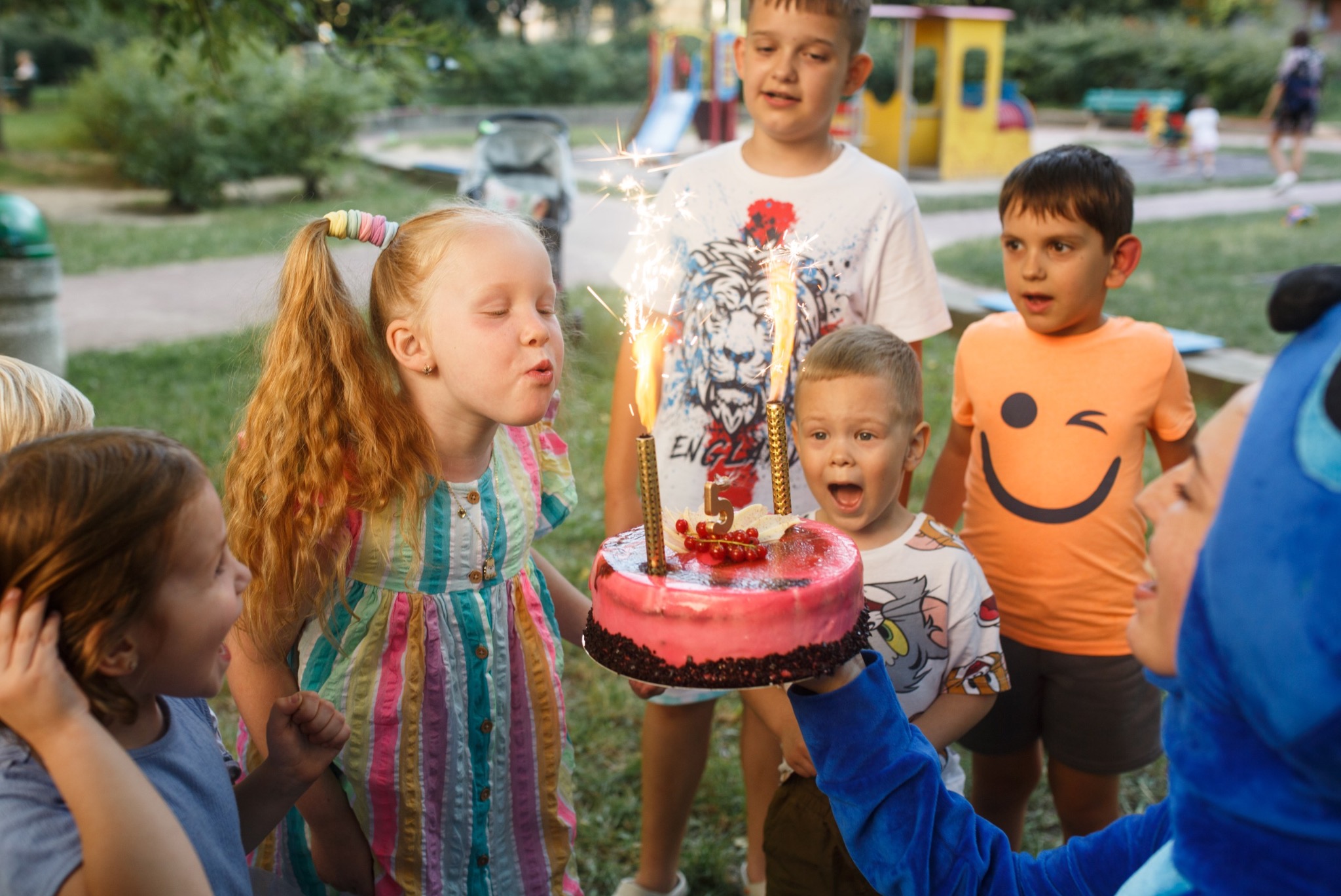 Urodziny dziecka w plenerze: dziewczynka zdmuchuje świeczki z tortu, otoczona przez animatora w kostiumie i grupę rozentuzjazmowanych dzieci.