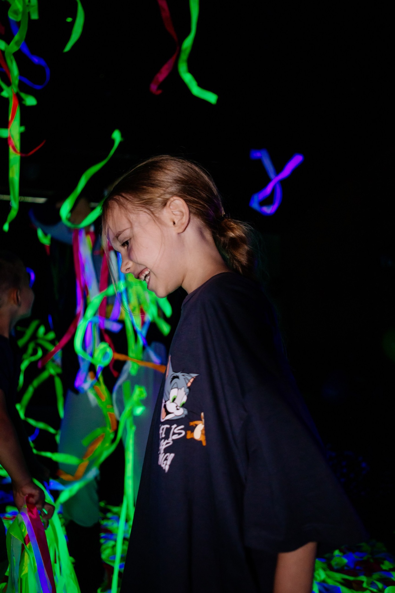 Uśmiechnięta dziewczynka w ciemnej koszulce z nadrukiem, otoczona neonowymi serpentynami podczas imprezy z animatorem. Dynamiczne, kolorowe oświetlenie.