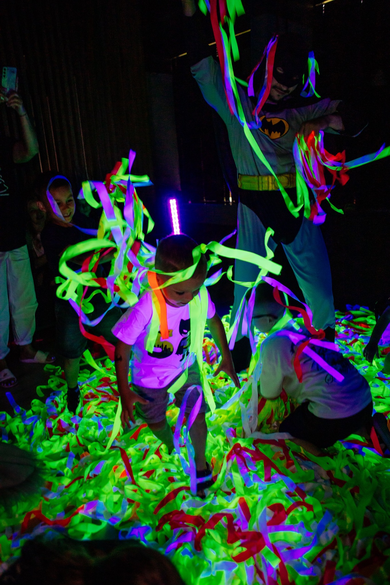 Uśmiechnięte dzieci bawią się w neonowych serpentynach pod czujnym okiem animatora przebranego za Batmana podczas imprezy urodzinowej w ciemnym pomieszczeniu.