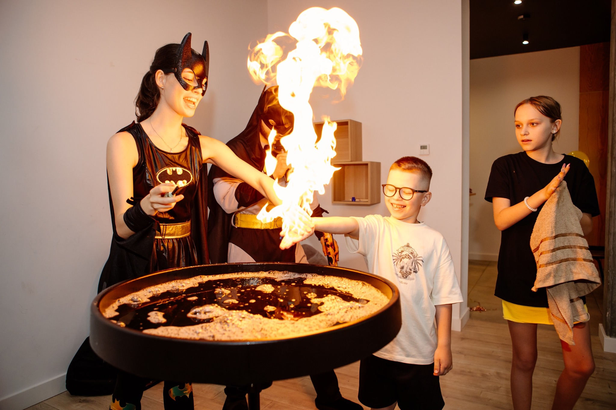 Animatorzy przebrani za Batmana i Batwoman prezentują pokaz iluzji z ogniem dla uśmiechniętego chłopca podczas imprezy urodzinowej. Dziewczynka z ręcznikiem obserwuje.