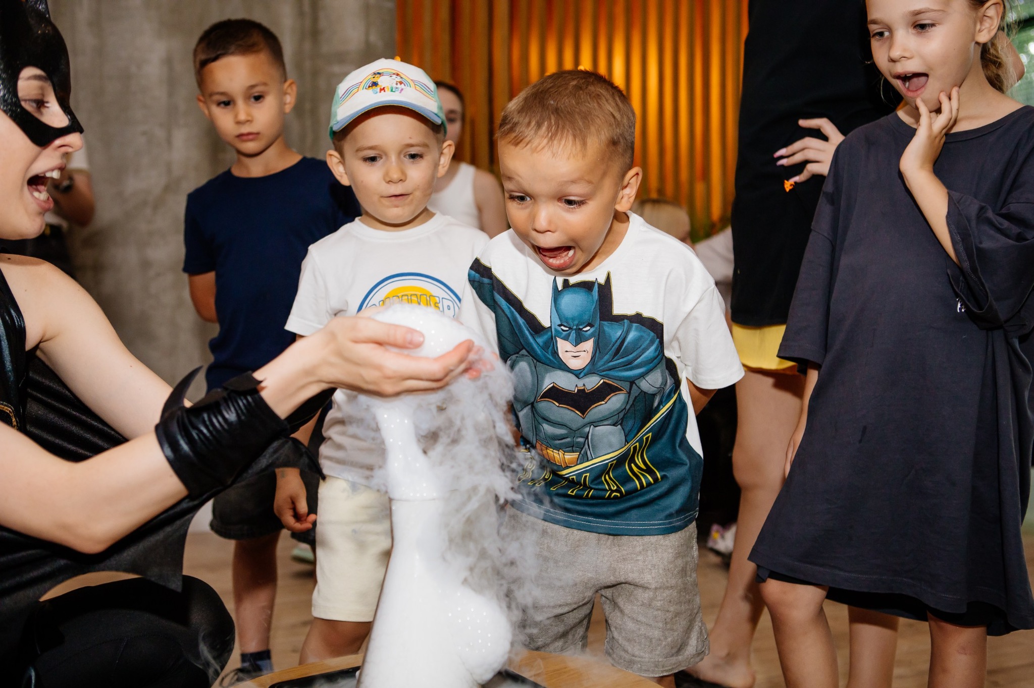 Animator w stroju Catwoman prezentuje eksperyment z suchym lodem grupie zaciekawionych dzieci podczas imprezy urodzinowej.