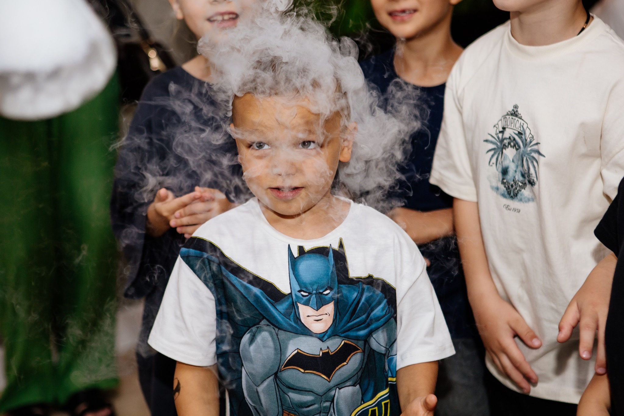 Chłopiec w koszulce z Batmanem otoczony dymem podczas pokazu dla dzieci. W tle widoczne inne dzieci i elementy dekoracji.