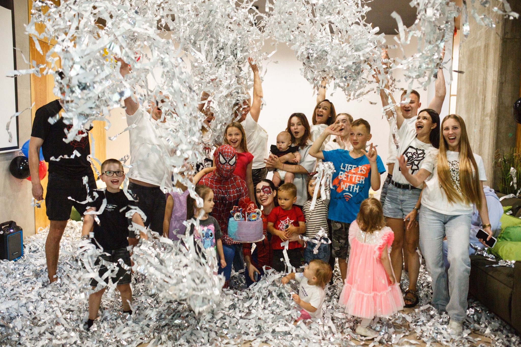 Radosne przyjęcie urodzinowe z animatorem w stroju Spider-Mana, tortem i deszczem srebrnego konfetti. Dzieci i dorośli świętują, uśmiechają się i bawią.