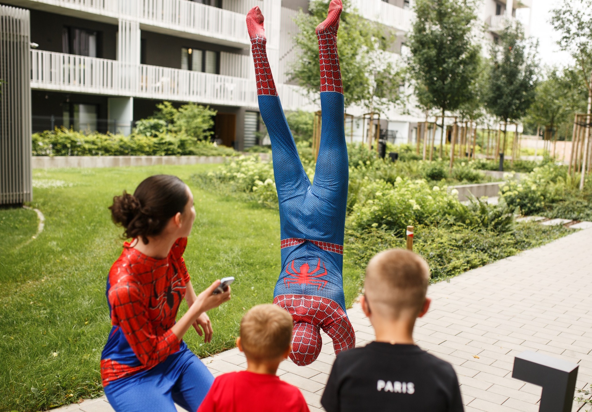 Animator w stroju Spider-Mana wykonuje akrobacje na trawniku, obserwują go dzieci i kobieta w stroju Spider-Girl. W tle budynek mieszkalny i zieleń.