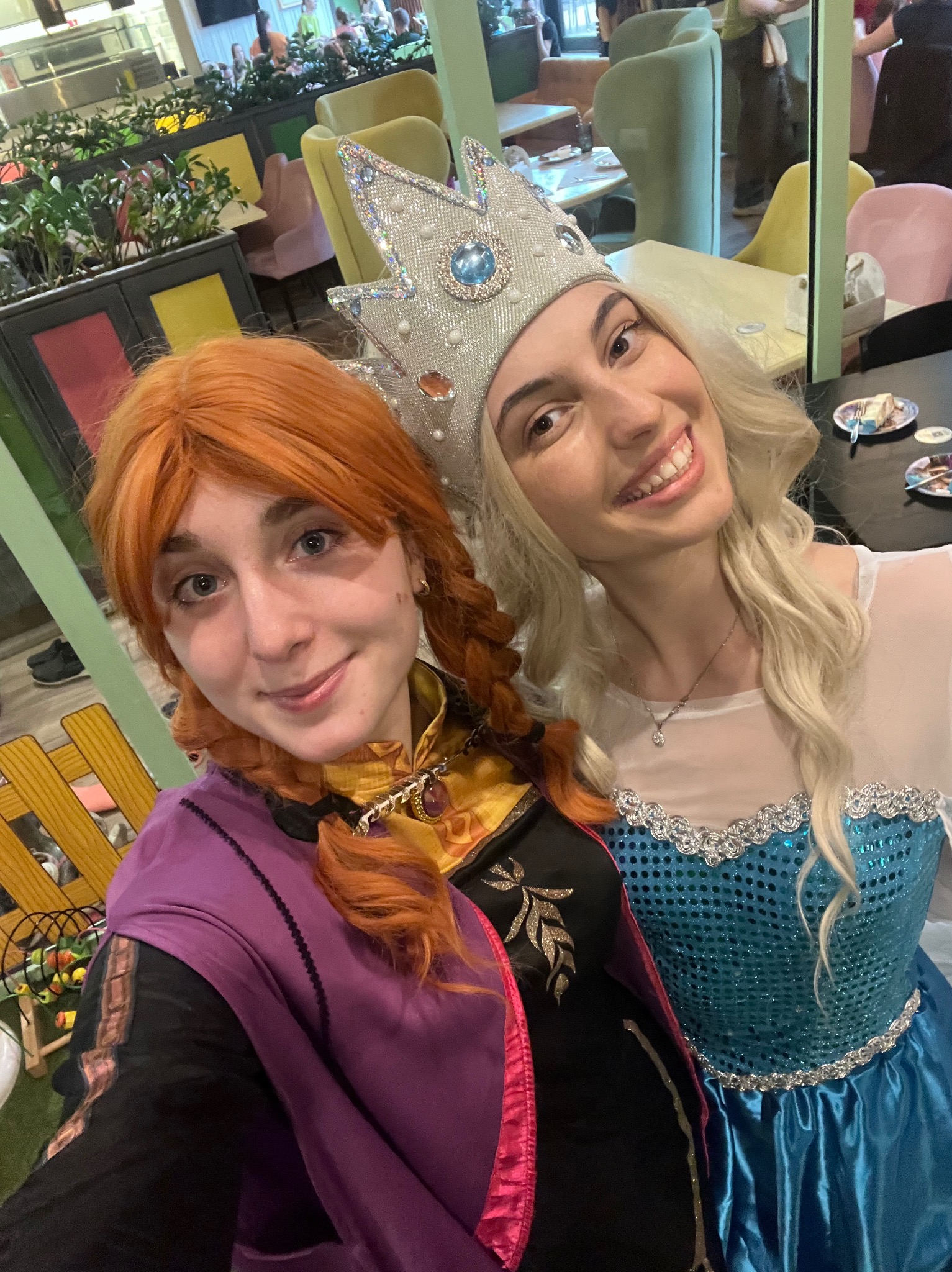 Uśmiechnięte animatorki w kostiumach Elsy i Anny z Krainy Lodu, pozują do selfie w kawiarni, widoczne dekoracje i stoliki.