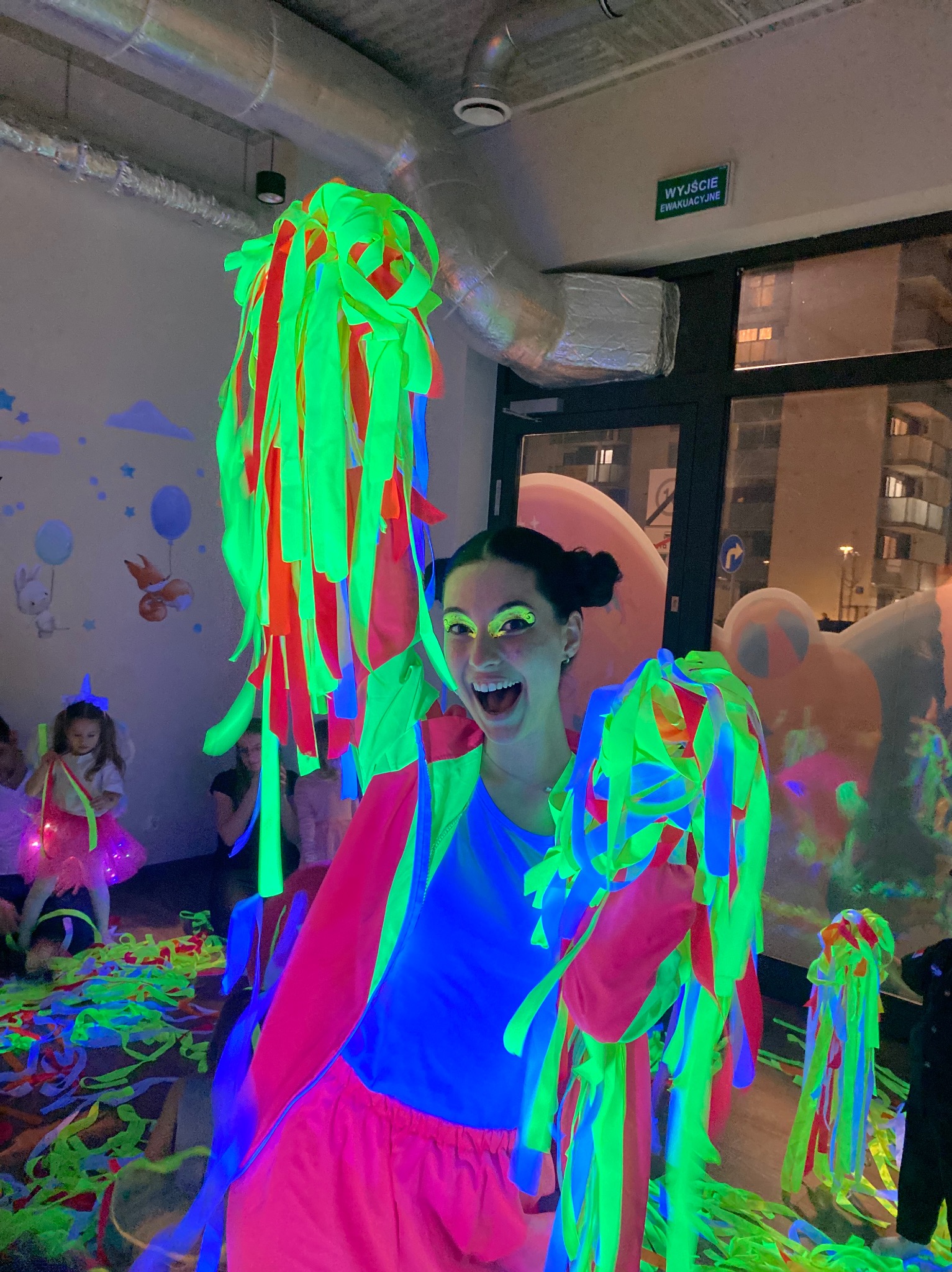 Uśmiechnięta animatorka w neonowym stroju, trzymająca kolorowe serpentyny, na tle dzieci i dekoracji w stylu UV party. Zabawa z animatorem dla dzieci.