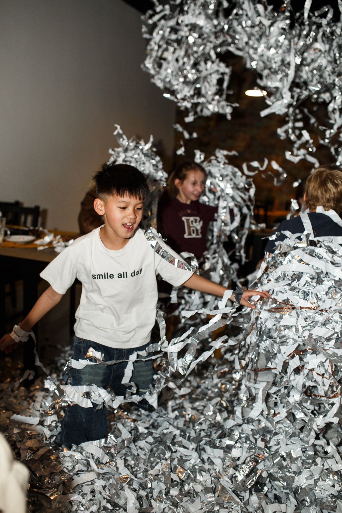 Radosny chłopiec w białej koszulce 'smile all day' bawi się z innymi dziećmi w srebrnych serpentynach podczas imprezy.