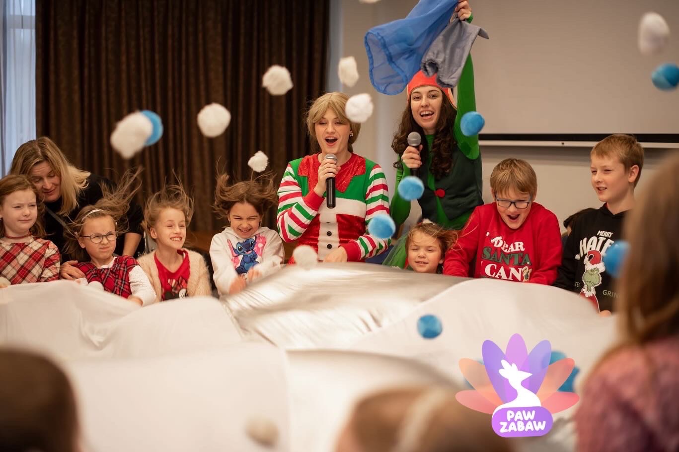 Animatorzy w strojach świątecznych prowadzą zabawy z dziećmi, rzucając miękkimi piłkami podczas imprezy z logo Paw Zabaw w tle.