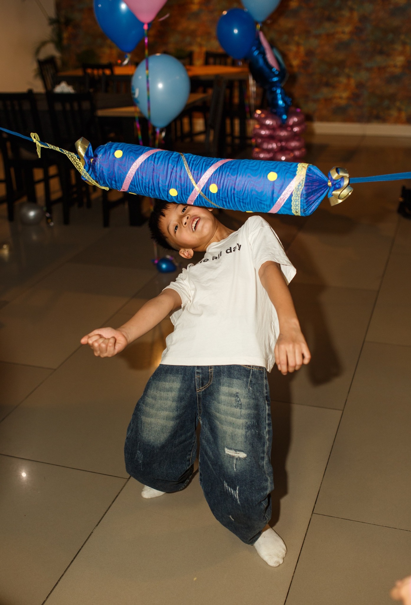 Chłopiec przechodzi pod kolorową poprzeczką limbo na tle sali z balonami, uśmiechając się podczas zabawy na przyjęciu urodzinowym.
