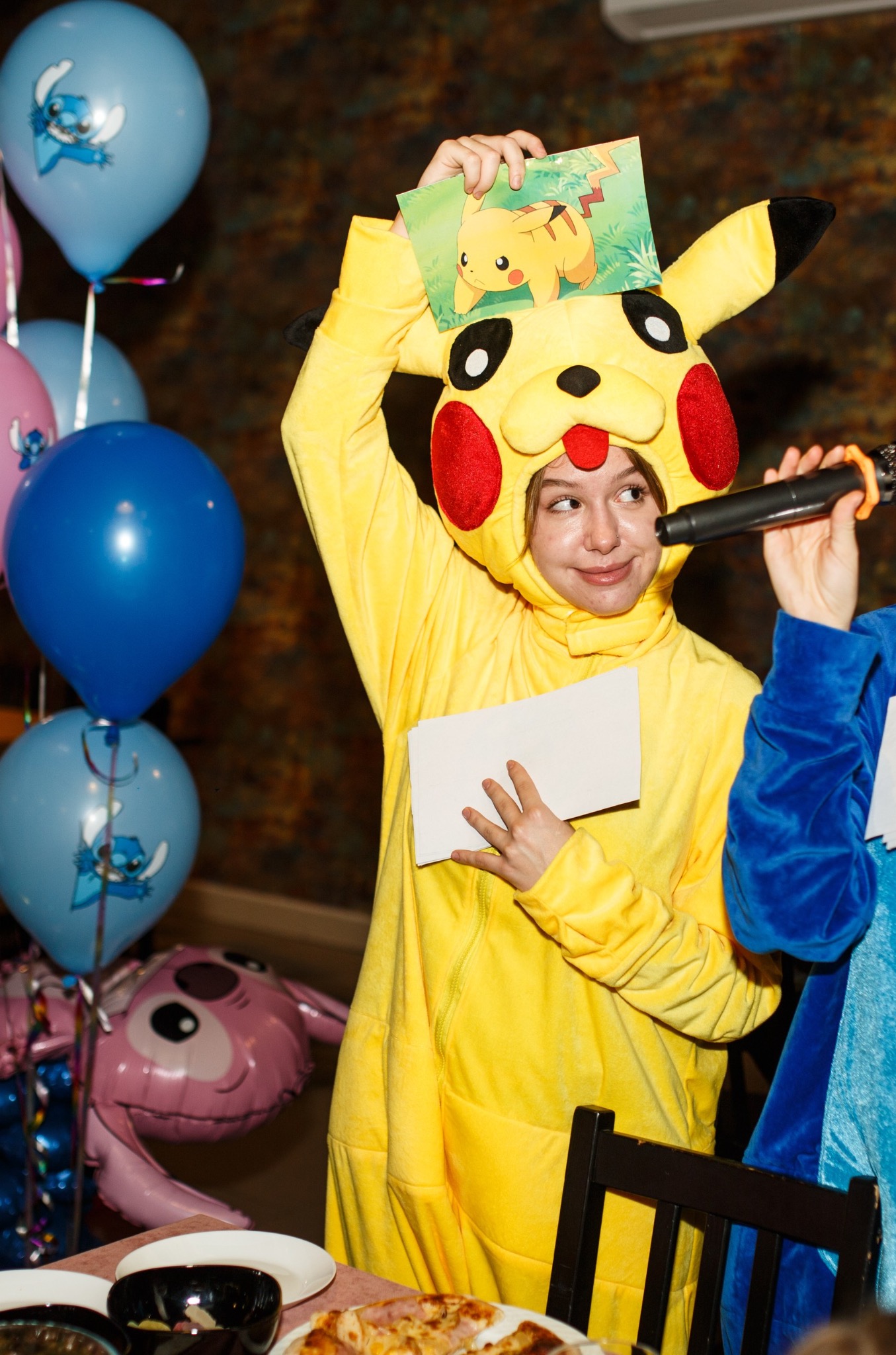 Osoba w kostiumie Pikachu trzyma kartkę z Pikachu i mikrofon, otoczona balonami z motywem Lilo i Stitch na imprezie urodzinowej.