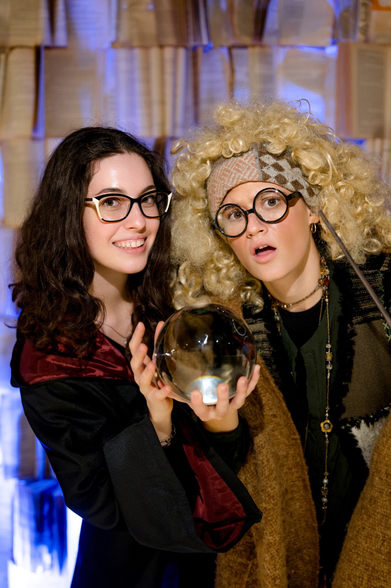 Dwie kobiety w kostiumach czarodziejek, trzymają kryształową kulę na tle dekoracji z książek. Tematyczna impreza, event dla dzieci.