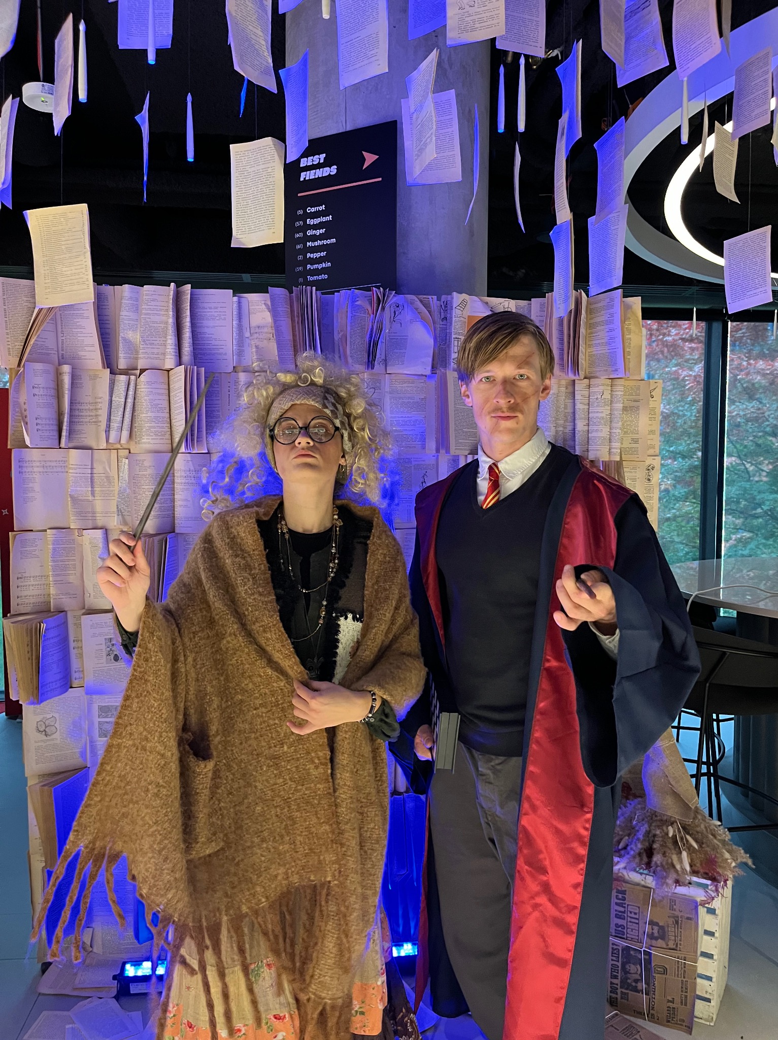 Dwie osoby w kostiumach tematycznych, kobieta z różdżką i mężczyzna w szacie, pozują na tle dekoracji z książek i świecących elementów, tworząc magiczną atmosferę.