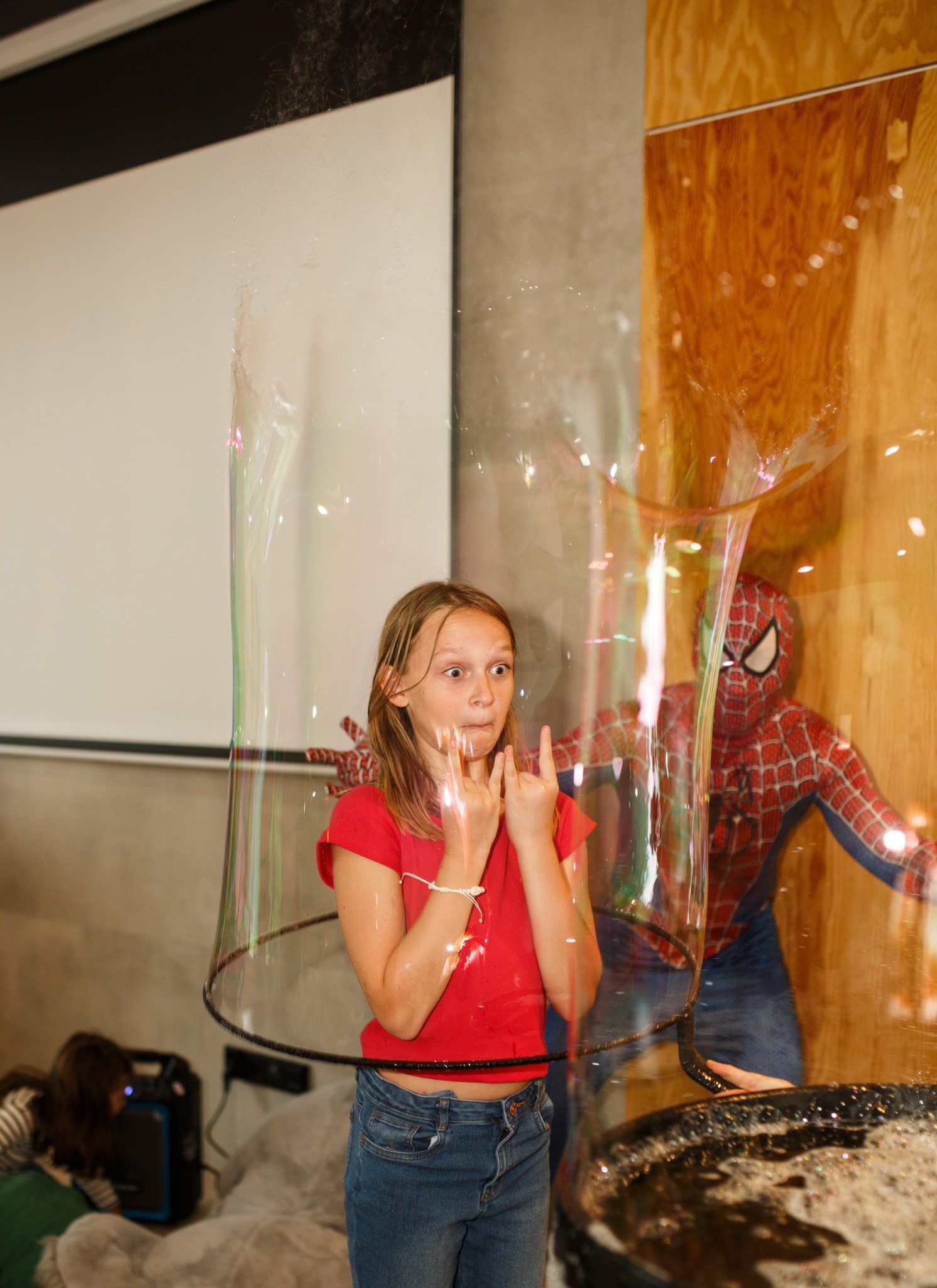 Dziewczynka w bańce mydlanej, w tle postać w kostiumie Spider-Mana. Atrakcja dla dzieci podczas imprezy, zabawa z animatorem i ogromnymi bańkami mydlanymi.