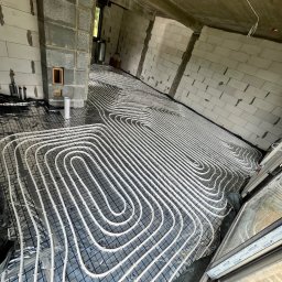 SAN-BUD CONSTRUCTIONS SPÓŁKA Z OGRANICZONĄ ODPOWIEDZIALNOŚCIĄ - Rozłożone białe rurki ogrzewania podłogowego na folii z siatką w surowym wnętrzu budynku z bloczków, widoczne okna i pion kanalizacyjny.