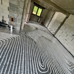 SAN-BUD CONSTRUCTIONS SPÓŁKA Z OGRANICZONĄ ODPOWIEDZIALNOŚCIĄ - Białe rury ogrzewania podłogowego ułożone na siatce w surowym wnętrzu budynku z betonowymi ścianami i oknem w tle.